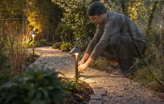 Person richtet eine niedrige warm leuchtende Gartenlampe am Kiesweg aus; Thema Japanischer Garten, Schwerpunkt Naturgarten; Ausgangslage.