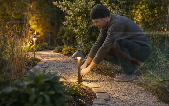 Person richtet eine niedrige warm leuchtende Gartenlampe am Kiesweg aus; Thema Japanischer Garten, Schwerpunkt Pflegeleichtigkeit; Ausgangslage.