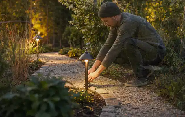 Person richtet eine niedrige warm leuchtende Gartenlampe am Kiesweg aus; Thema Japanischer Garten, Schwerpunkt Privatsphäre; Praxisalltag.
