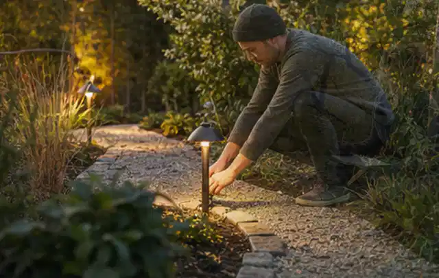 Person richtet eine niedrige warm leuchtende Gartenlampe am Kiesweg aus; Thema Kleingarten, Schwerpunkt Anleitung; Umsetzung.