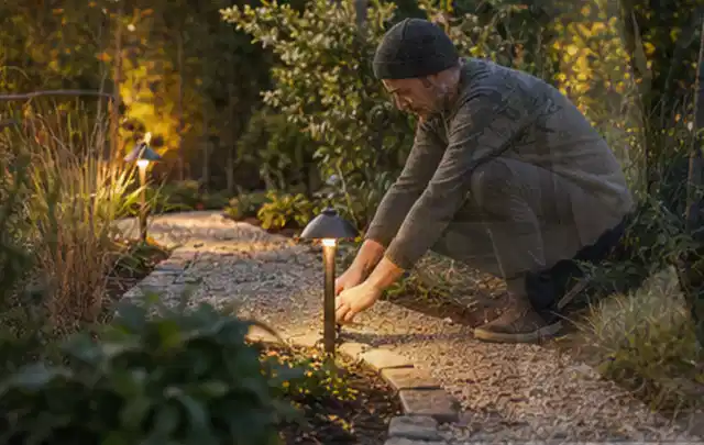 Person richtet eine niedrige warm leuchtende Gartenlampe am Kiesweg aus; Thema Kleingarten, Schwerpunkt Checkliste; Kontrolle.