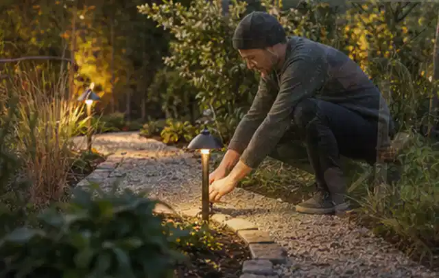 Person richtet eine niedrige warm leuchtende Gartenlampe am Kiesweg aus; Thema Kleingarten, Schwerpunkt Fehlerhilfe; Ergebnis.
