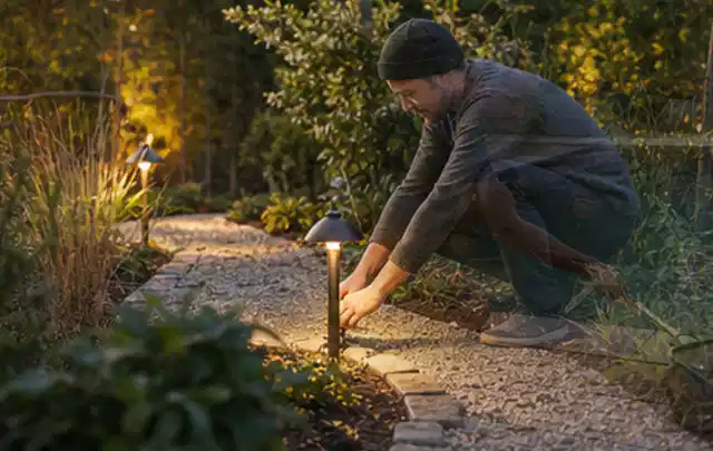 Person richtet eine niedrige warm leuchtende Gartenlampe am Kiesweg aus; Thema Kleingarten, Schwerpunkt Gärten mit kleiner Fläche; Umsetzung.