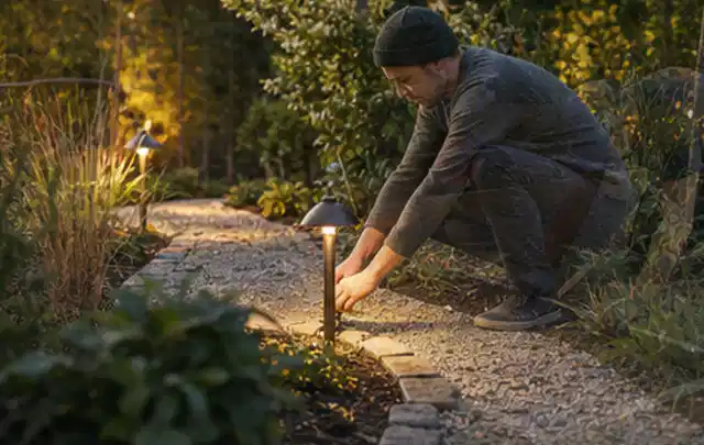 Person richtet eine niedrige warm leuchtende Gartenlampe am Kiesweg aus; Thema Kleingarten, Schwerpunkt Kosten; Kontrolle.
