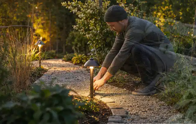 Person richtet eine niedrige warm leuchtende Gartenlampe am Kiesweg aus; Thema Kleingarten, Schwerpunkt Pflegeleichtigkeit; Umsetzung.