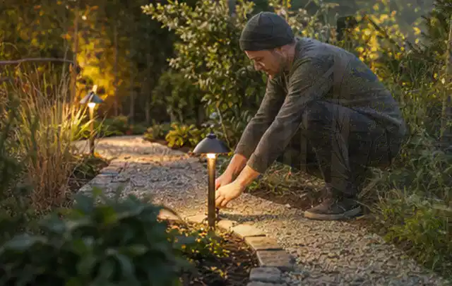 Person richtet eine niedrige warm leuchtende Gartenlampe am Kiesweg aus; Thema Kleingarten, Schwerpunkt Privatsphäre; Ergebnis.
