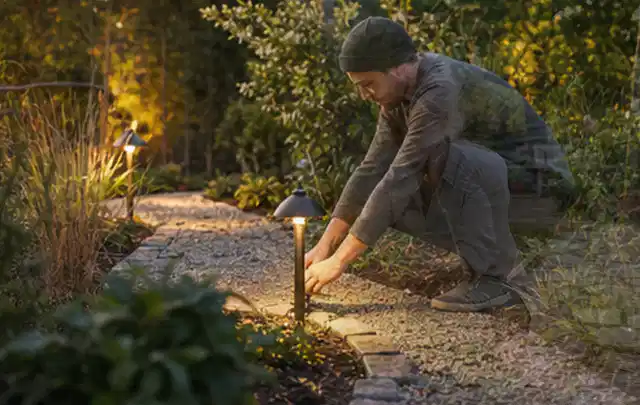 Person richtet eine niedrige warm leuchtende Gartenlampe am Kiesweg aus; Thema Kleingarten, Schwerpunkt Wegeführung; Praxisalltag.