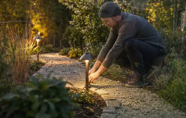 Person richtet eine niedrige warm leuchtende Gartenlampe am Kiesweg aus; Thema Kress Mission, Schwerpunkt Bestenliste; Ausgangslage.
