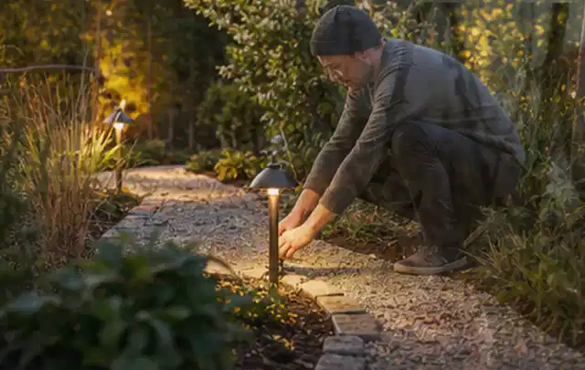 Person richtet eine niedrige warm leuchtende Gartenlampe am Kiesweg aus; Thema Kress Mission, Schwerpunkt Dimensionierung; Ausgangslage.