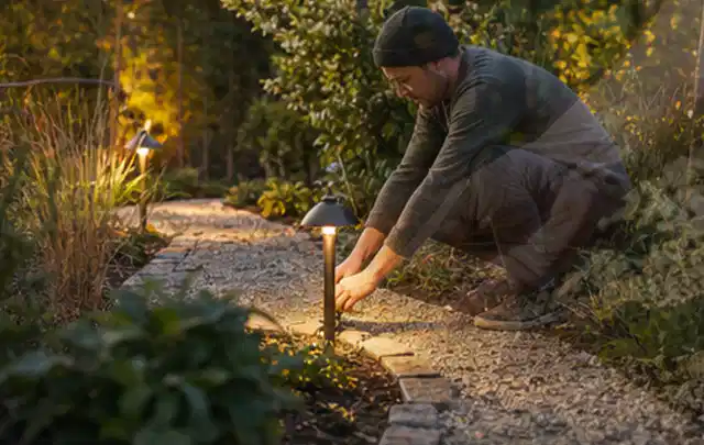 Person richtet eine niedrige warm leuchtende Gartenlampe am Kiesweg aus; Thema Kress Mission, Schwerpunkt Ersatzteile; Kontrolle.