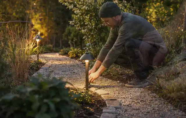 Person richtet eine niedrige warm leuchtende Gartenlampe am Kiesweg aus; Thema Kress Mission, Schwerpunkt Mähroboter; Ergebnis.