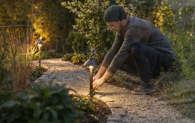 Person richtet eine niedrige warm leuchtende Gartenlampe am Kiesweg aus; Thema Kress Mission, Schwerpunkt Reinigung; Umsetzung.