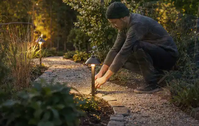 Person richtet eine niedrige warm leuchtende Gartenlampe am Kiesweg aus; Thema Kress Mission, Schwerpunkt Systemvergleich; Ausgangslage.