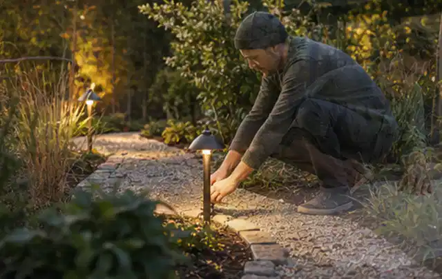 Person richtet eine niedrige warm leuchtende Gartenlampe am Kiesweg aus; Thema Kress Mission, Schwerpunkt Vergleich; Umsetzung.