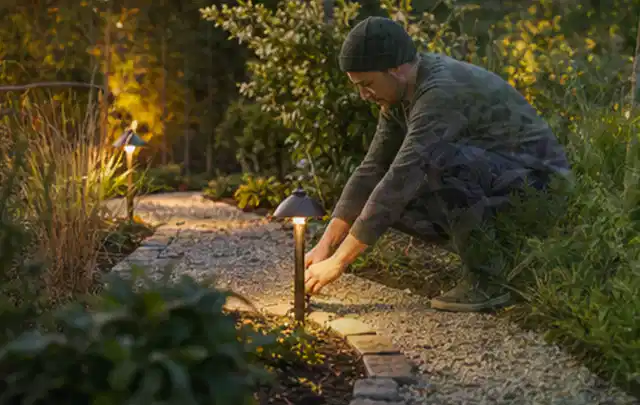 Person richtet eine niedrige warm leuchtende Gartenlampe am Kiesweg aus; Thema Kress Mission, Schwerpunkt Winterlager; Kontrolle.