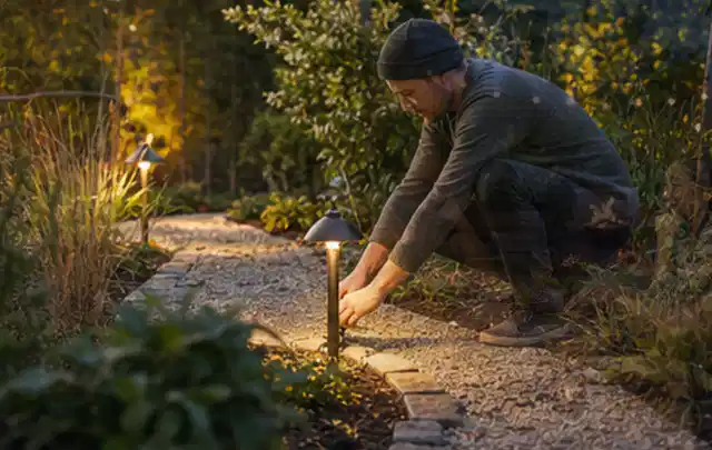 Person richtet eine niedrige warm leuchtende Gartenlampe am Kiesweg aus; Thema Lagerhaus im Garten, Schwerpunkt Aufbau; Ausgangslage.