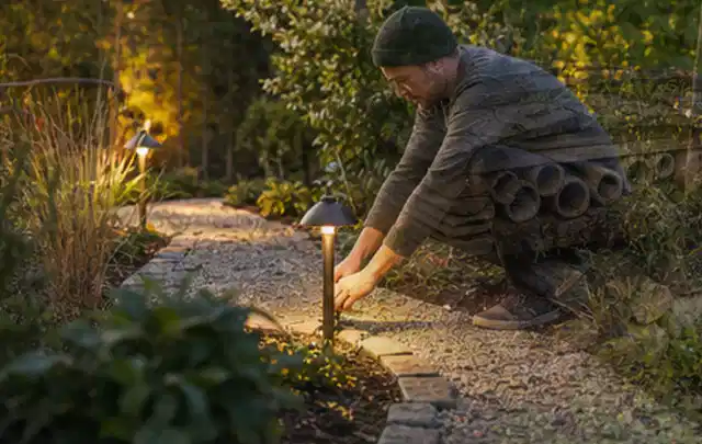Person richtet eine niedrige warm leuchtende Gartenlampe am Kiesweg aus; Thema Lagerhaus im Garten, Schwerpunkt Baurecht; Ausgangslage.