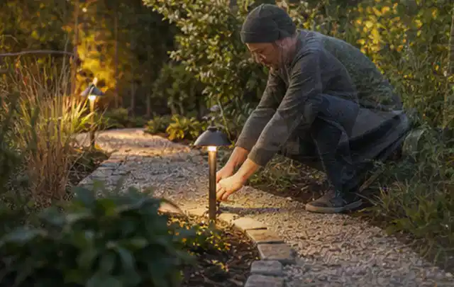 Person richtet eine niedrige warm leuchtende Gartenlampe am Kiesweg aus; Thema Lagerhaus im Garten, Schwerpunkt DIY; Umsetzung.