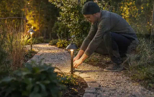 Person richtet eine niedrige warm leuchtende Gartenlampe am Kiesweg aus; Thema Lagerhaus im Garten, Schwerpunkt Fehlerhilfe; Ausgangslage.