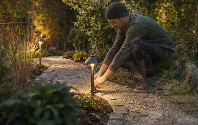 Person richtet eine niedrige warm leuchtende Gartenlampe am Kiesweg aus; Thema Lagerhaus im Garten, Schwerpunkt Winterschutz; Ausgangslage.