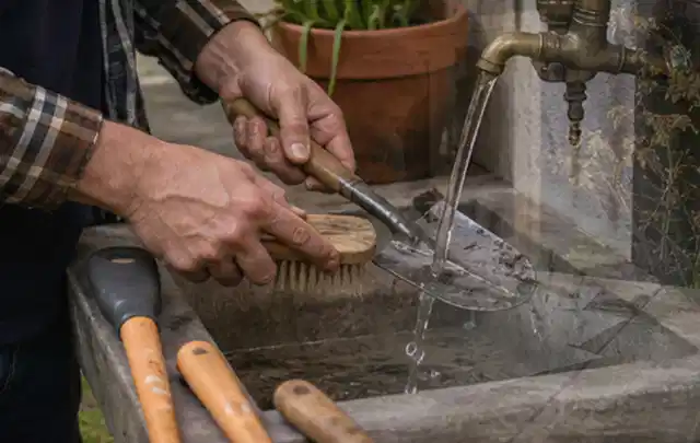 Hände reinigen gebrauchte Gartengeräte mit Bürste und Wasser am Außenbecken; Thema Mähroboter Akkus, Schwerpunkt Anwendung; Praxisalltag.