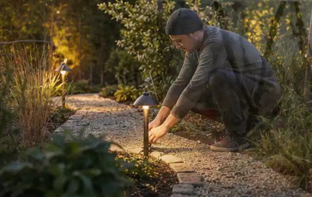 Person richtet eine niedrige warm leuchtende Gartenlampe am Kiesweg aus; Thema Mähroboter Akkus, Schwerpunkt Bestenliste; Praxisalltag.