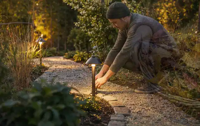 Person richtet eine niedrige warm leuchtende Gartenlampe am Kiesweg aus; Thema Mähroboter Akkus, Schwerpunkt Fehlerhilfe; Ergebnis.