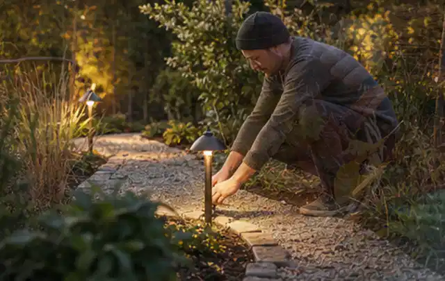 Person richtet eine niedrige warm leuchtende Gartenlampe am Kiesweg aus; Thema Mähroboter Akkus, Schwerpunkt Mähroboter; Kontrolle.