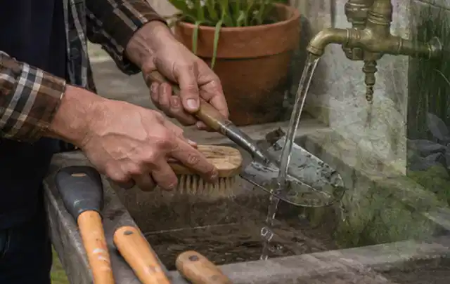 Hände reinigen gebrauchte Gartengeräte mit Bürste und Wasser am Außenbecken; Thema Mähroboter Akkus, Schwerpunkt Mähroboter; Ergebnis.