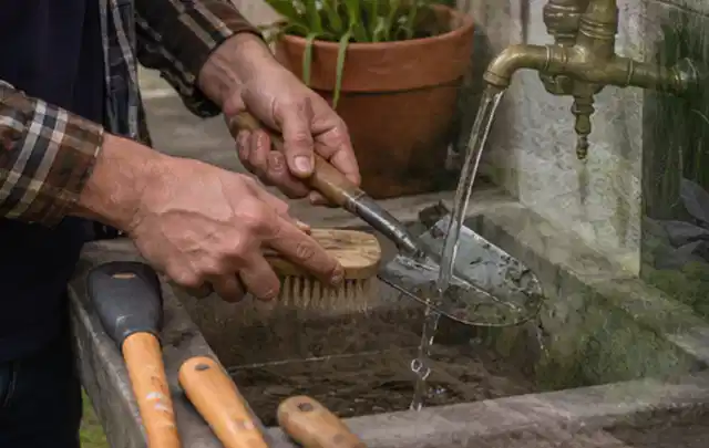 Hände reinigen gebrauchte Gartengeräte mit Bürste und Wasser am Außenbecken; Thema Mähroboter Akkus, Schwerpunkt Sicherheit; Kontrolle.