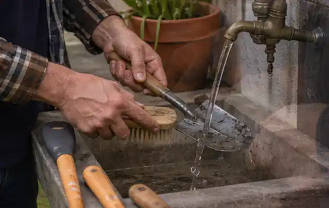 Hände reinigen gebrauchte Gartengeräte mit Bürste und Wasser am Außenbecken; Thema Mähroboter Akkus, Schwerpunkt Vergleich; Praxisalltag.
