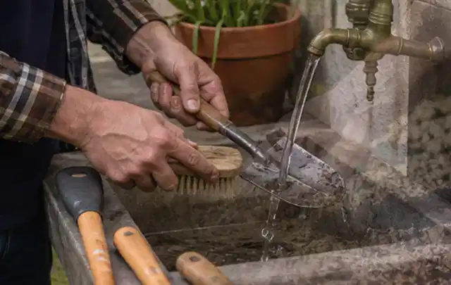 Hände reinigen gebrauchte Gartengeräte mit Bürste und Wasser am Außenbecken; Thema Mähroboter Akkus, Schwerpunkt Winterlager; Umsetzung.