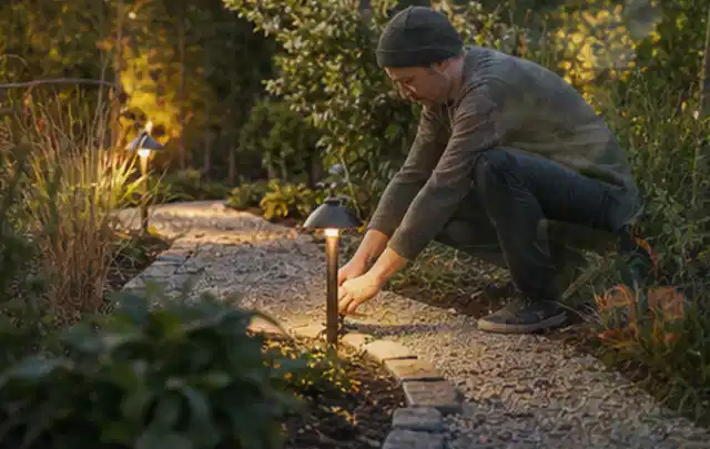 Person richtet eine niedrige warm leuchtende Gartenlampe am Kiesweg aus; Thema Mähroboter Akkus, Schwerpunkt Winterlager; Kontrolle.
