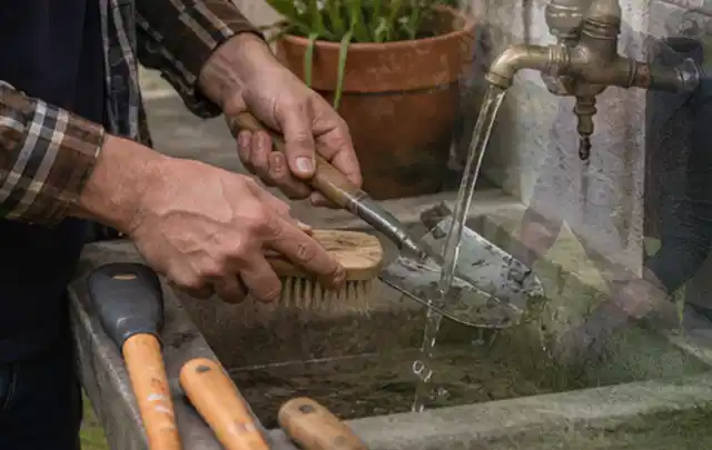 Hände reinigen gebrauchte Gartengeräte mit Bürste und Wasser am Außenbecken; Thema Mähroboter für große Gärten, Schwerpunkt Bestenliste; Umsetzung.