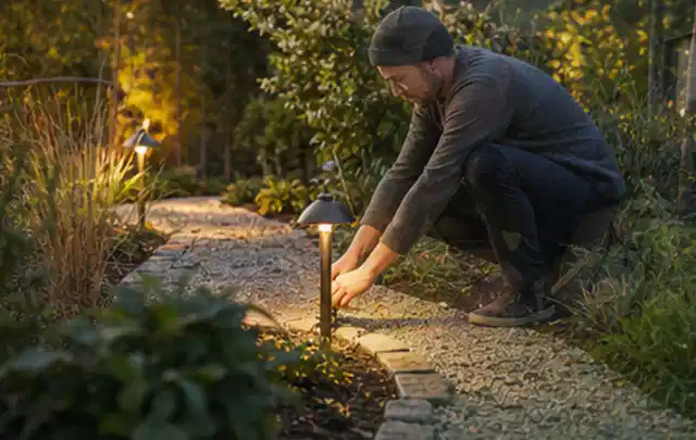 Person richtet eine niedrige warm leuchtende Gartenlampe am Kiesweg aus; Thema Mähroboter für große Gärten, Schwerpunkt Kosten; Umsetzung.