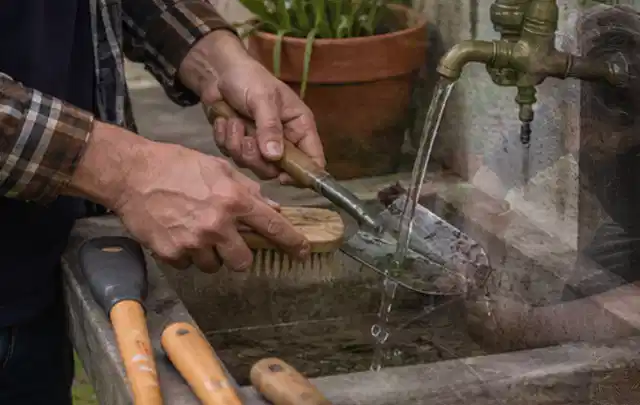 Hände reinigen gebrauchte Gartengeräte mit Bürste und Wasser am Außenbecken; Thema Mähroboter für große Gärten, Schwerpunkt Leistung; Praxisalltag.