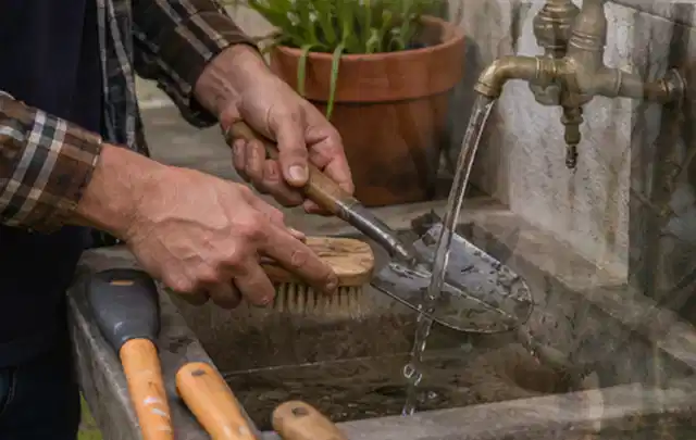 Hände reinigen gebrauchte Gartengeräte mit Bürste und Wasser am Außenbecken; Thema Mähroboter für große Gärten, Schwerpunkt Nachhaltigkeit; Umsetzung.