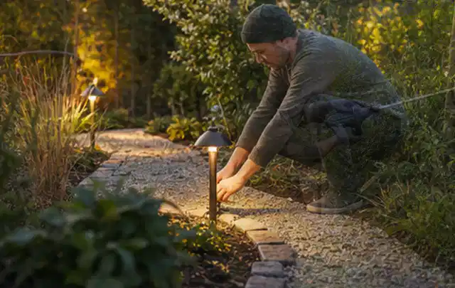 Person richtet eine niedrige warm leuchtende Gartenlampe am Kiesweg aus; Thema Mähroboter für große Gärten, Schwerpunkt Testkriterien; Ausgangslage.