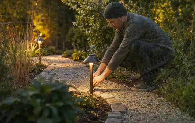 Person richtet eine niedrige warm leuchtende Gartenlampe am Kiesweg aus; Thema Mähroboter für große Gärten, Schwerpunkt Wartung; Kontrolle.