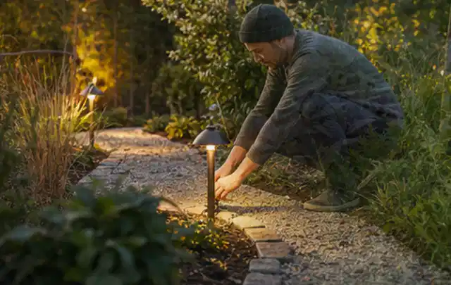 Person richtet eine niedrige warm leuchtende Gartenlampe am Kiesweg aus; Thema Mähroboter für Hanglagen, Schwerpunkt Dimensionierung; Ausgangslage.