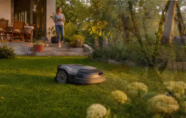 Mähroboter fährt am Abend über einen Rasen im Familiengarten; Thema Mähroboter für Hanglagen, Schwerpunkt Ersatzteile; Kontrolle.