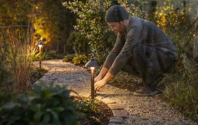 Person richtet eine niedrige warm leuchtende Gartenlampe am Kiesweg aus; Thema Mähroboter für Hanglagen, Schwerpunkt Fehlerhilfe; Ausgangslage.