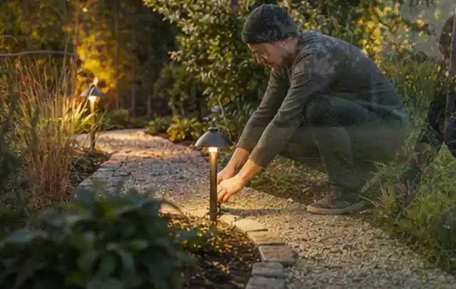 Person richtet eine niedrige warm leuchtende Gartenlampe am Kiesweg aus; Thema Mähroboter für Hanglagen, Schwerpunkt Nachhaltigkeit; Kontrolle.