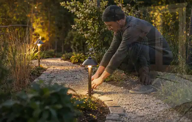 Person richtet eine niedrige warm leuchtende Gartenlampe am Kiesweg aus; Thema Mähroboter für Hanglagen, Schwerpunkt Reinigung; Kontrolle.
