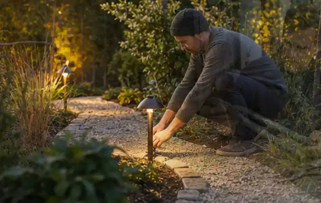 Person richtet eine niedrige warm leuchtende Gartenlampe am Kiesweg aus; Thema Mähroboter für Hanglagen, Schwerpunkt Reparatur; Umsetzung.