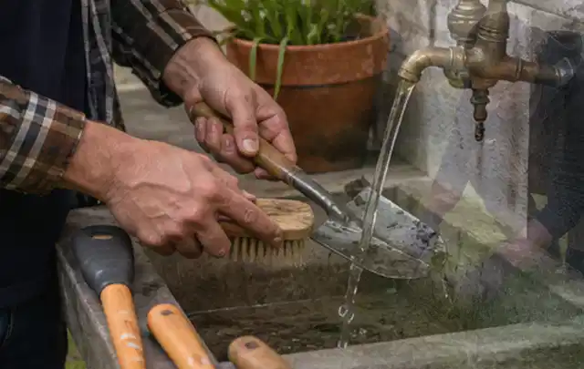Hände reinigen gebrauchte Gartengeräte mit Bürste und Wasser am Außenbecken; Thema Mähroboter für Hanglagen, Schwerpunkt Reparatur; Ergebnis.