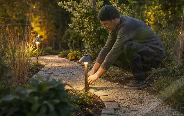Person richtet eine niedrige warm leuchtende Gartenlampe am Kiesweg aus; Thema Mähroboter für Hanglagen, Schwerpunkt Sicherheit; Ergebnis.