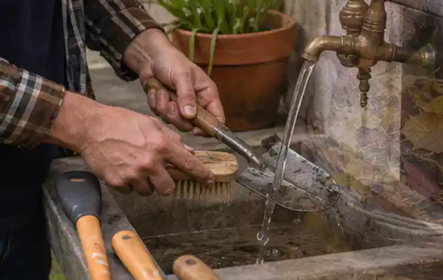 Hände reinigen gebrauchte Gartengeräte mit Bürste und Wasser am Außenbecken; Thema Mähroboter für Hanglagen, Schwerpunkt Systemvergleich; Praxisalltag.