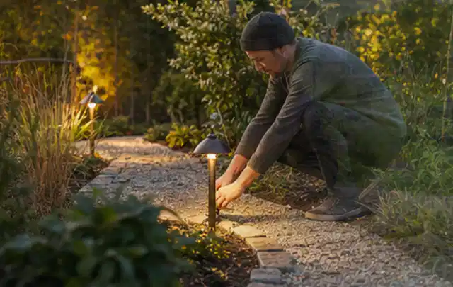 Person richtet eine niedrige warm leuchtende Gartenlampe am Kiesweg aus; Thema Mähroboter für Hanglagen, Schwerpunkt Testkriterien; Umsetzung.