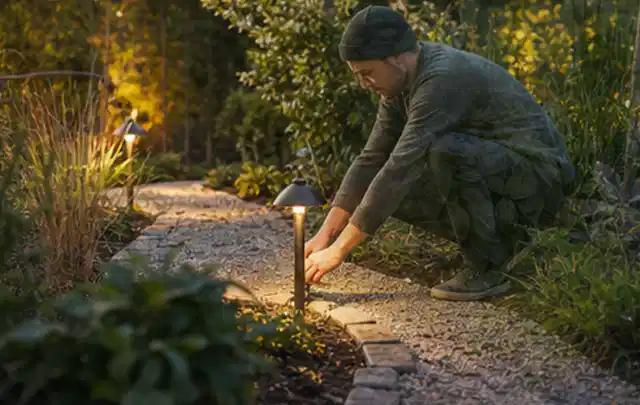 Person richtet eine niedrige warm leuchtende Gartenlampe am Kiesweg aus; Thema Mähroboter für kleine Gärten, Schwerpunkt Dimensionierung; Kontrolle.